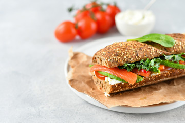 sandwich from a cereal baguette with avocado, salmon, cream cheese, tomatoes and lettuce leaves on a white plate.  light background, selective focus and copy space