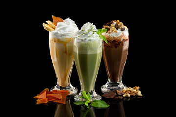 Three glasses with caramel, mint and chocolate cocktails decorated with whipped cream at wooden board isolated at black background.