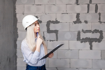 Closeup blonde girl foreman in white construction helmet holding pen, folder with plan in house under construction. Concept professional designer plans project, measures the room, designs walls