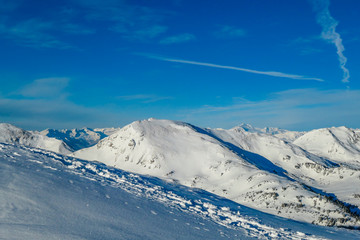 A beautiful and serene landscape of mountains covered with snow. Thick snow covers the slopes. Few plane tracks on the sky. Clear weather. Powder snow with a path marked by people walking by. 