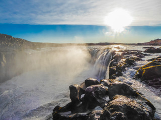 A waterfall splashing a lot of water drops around, caught on a sunny day. Lots of sharp rocks around the waterfall. Wide bank of the river. Amazing power and beauty of nature. Untouched landscape.
