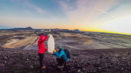 A couple standing on a lava field lights up a wish lantern. Girl is holding the lantern, boy is lighting it up. In the back sky is exploding with colors of sunrise. Harsh and barren landscape. © Chris