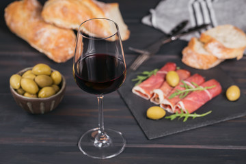 Glass of red wine with slices of cured ham or Spanish jamon serrano or Italian prosciutto crudo, bread, green olives and arugula