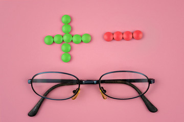 Glasses with plus and minus made of green and red pills on pink background. Concept of eyesight...