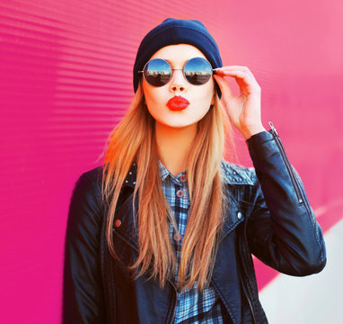 Fashion portrait beautiful blonde woman sending sweet air kiss in rock black style jacket, hat on city street over colorful pink wall background