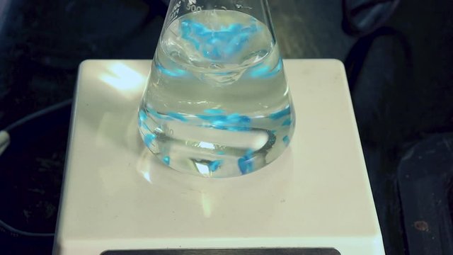 Close up of blue crystals of copper sulfate pentahydrate being added to erlenmeyer flask on magnetic stirrer