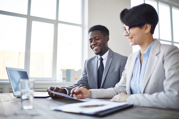 Two confident and successful colleagues in formalwear discussing financial paper by workplace in...