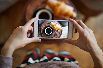Overview of picture of cup of tea or coffee and fresh croissant in smartphone held by young woman
