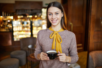 Pretty casual girl with toothy smile holding black cup with tea or coffee while having rest in cafeteria