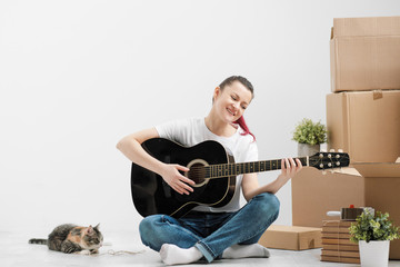 A young woman in a white T-shirt is resting and playing the guitar on the background of cardboard boxes in a new empty apartment with little pet cat.