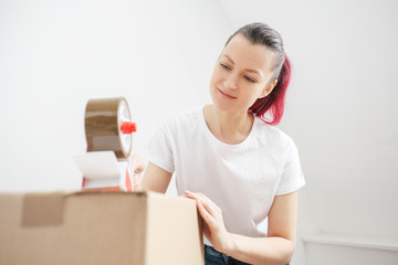 Young beautiful brunette girl in a white t-shirt packs cardboard boxes with a dispenser and adhesive scotch adhesive tape. The concept of moving to a new home.