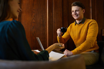 Happy and successful casual young businessman with cup of tea talking to his colleague in cafe