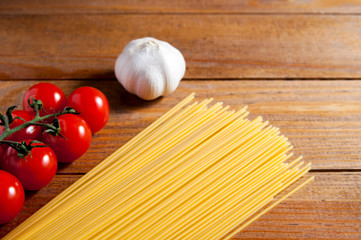 Spaghetti, cherry tomatoes and bulb of garlic on a brown wooden table. Tomatoes and garlic to the left of spaghetti.