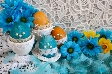 Wielkanocna kompozycja z niebieskimi pisankami, żółtymi i niebieskimi kwiatami oraz piórami na tle białej koronki © Monika