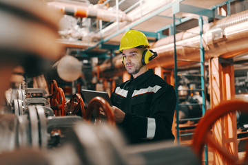 Young Caucasian hardworking heavy industry worker in protective suit and helmet using tablet and...