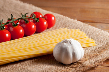 Spaghetti, cherry tomatoes and bulb of garlic on burlap on a brown wooden table. Tomatoes to the left of spaghetti, garlic to the right. Close-up.