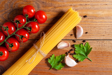 Bunch of raw spaghetti tied with rope, tomatoes cherry, slices of garlic, parsley leaves and pepper on brown wooden background. Top view.
