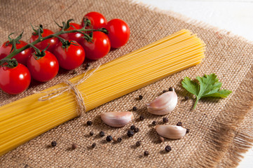 Bunch of raw spaghetti, cherry tomatoes, peppers, garlic cloves and parsley leaf on burlap on a white wooden background. Close-up.