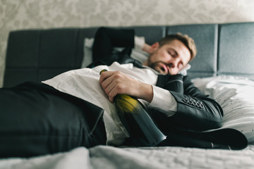 Drunk businessman in suit lying in the bed and sleeping with bottle of alcohol in his hand. Alcohol abuse concept.