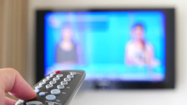 An interior shot focused on a remote control switching channels on a blurred television screen.