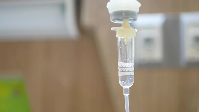 Saline drop in a drip chamber being infused along with medicine, and nutrients into a circulatory system of a patient, with a defocus of hospital patient room in the background