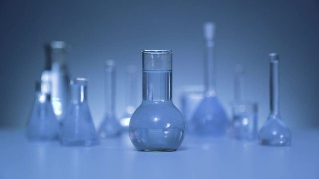 On the table is a medical flask with liquid. The camera smoothly leaves the flask. Gray wall on background. Flasks in defocus on the gray background.