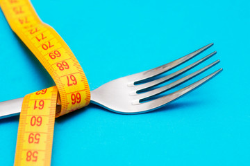 Tape measure around a fork as concept for diet. Fork are wrapped in yellow measuring tape on blue background
