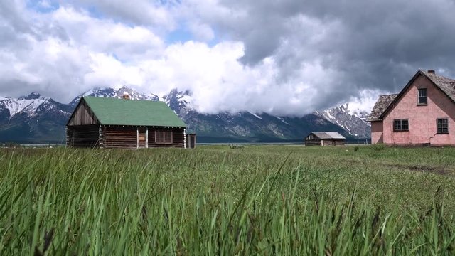 Log cabin in green grass in front of snowy mountains with dark clouds at Moulton Barn on Mormon Row in Grand Teton National Park, Wyoming