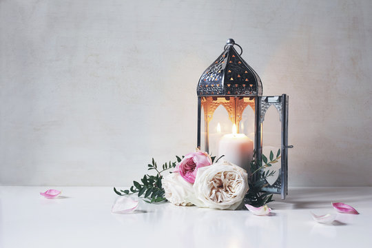 Vintage silver Moroccan, Arabic lantern with glowing candle, green branches, rose flowers and pink petals on white table background. Greeting card for Muslim holiday Ramadan Kareem. Shaby wall.