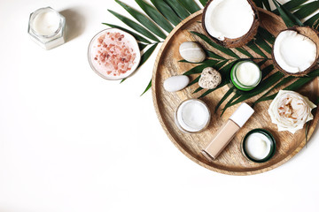 Fototapeta na wymiar Styled beauty composition. Skin creams, makeup bottle, rose and pebble stones on wooden tray. Coconuts, tropical palm leaves decoration. Cosmetics, spa concept. Empty space, flat lay, top view.