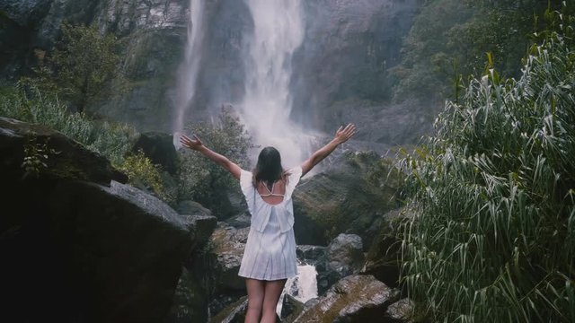 Back view happy young tourist woman with arms wide open enjoying moments of freedom at epic Sri Lanka jungle waterfall.