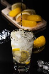 Pouring Sparkling cold water detox with lemon slices and ice on a dark glass background with black back