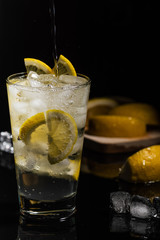 Pouring Sparkling water or other transparent drink with a lemons slices and ice cubes in Beautiful glass on black background horizontal view