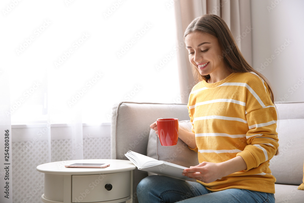 Wall mural Young woman with cup of coffee reading book on couch at home