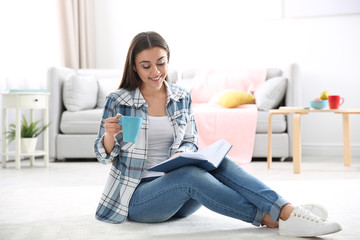Beautiful young woman with cup of coffee reading book on floor at home