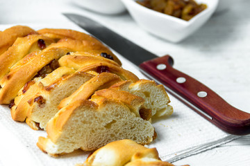 Piece of Sweet Braided Bread with raisins and knife on kitchen towel on white wooden background.