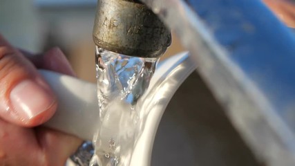 Hand washing white cup with a washcloth under a flow of water outdoors in summer