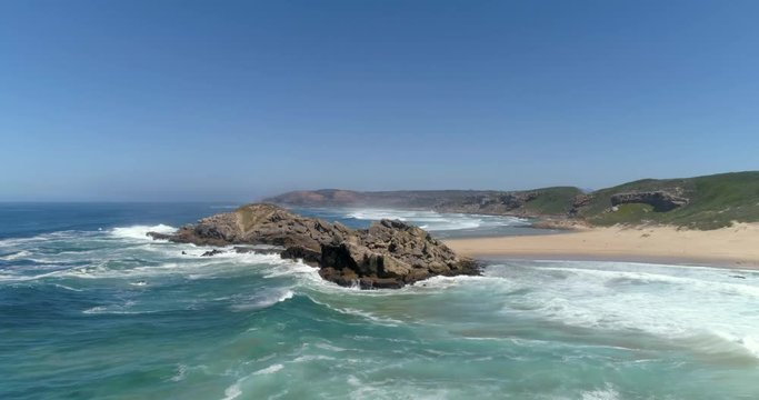 Aerial moving over Robberg Nature Reserve island, clear blue skies and white sandy beach, with wave and seagull action.