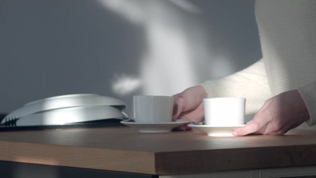Someone picks up two coffee settings near an open kitchen window as the curtain blows in the foreground.