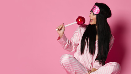 Beautiful young brunette woman in pajama hold huge sweet lollypop on pink background 