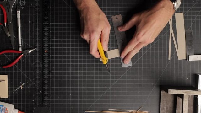 Overhead view of a male architect's hands scoring a strip of cardboard and folding it into a box shape. Shot has a black cutting mat as the background.