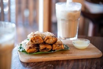 Fried banana with cheese topping on the top of banana leaf and wooden cutting board with blur glass of chocolate milk on background near the windows of coffee shop. Selective focus.