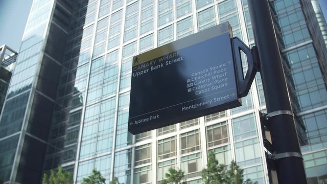 Low angle shot of a Canary Wharf sign in the street with big glass buildings behind.
