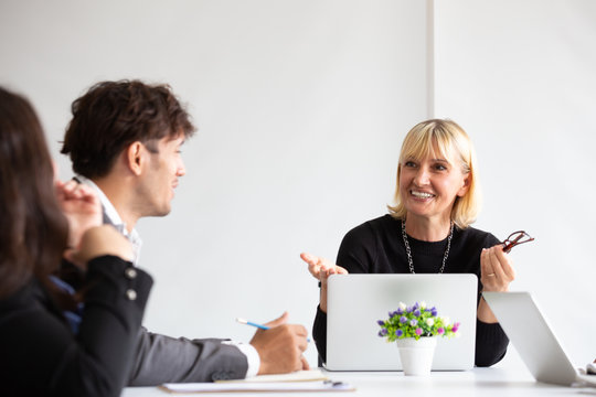 Smiling aged business woman talking and looking at colleague while team meeting, Happy attentive female team leader and coach listening and developing for new project idea