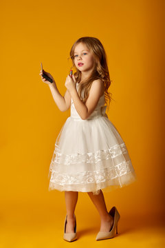 Adorable little girl in oversized shoes holding pocket mirror and applying lipstick while standing on yellow background