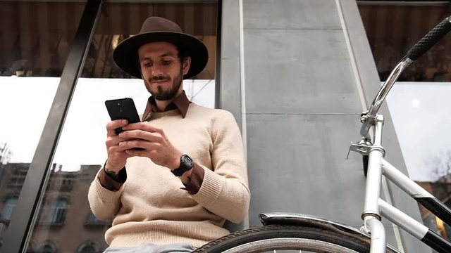 Stylish young bearded man with bicycle surfing internet on smartphone
