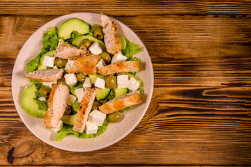Fresh salad with chicken meat, feta cheese, avocado, green olives and lettuce leaves in ceramic plate on wooden table. Top view