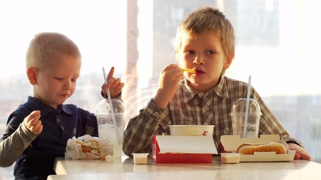 Children have lunch in fast food cafe. Boys drink lemonade and eat hamburgers in background of parking lot and cityscape. Blond kid are happy