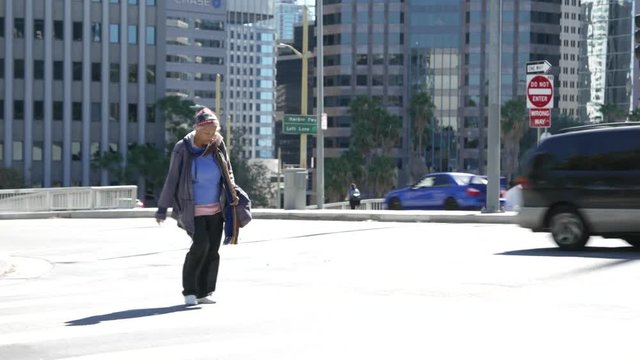 Homeless woman crosses the street with homeless encampment in background.