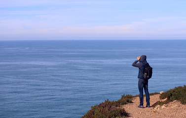 A man with a backpack looks through binoculars into the distance of the ocean. Cabo da Roca, Portugal. Cropped shot, horizontal, side view, perspective view. Concept of hobby and rest.